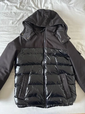 Svart pufferjacka från Calvin Klein - Snygg svart pufferjacka från Calvin Klein med glansig vadderad framsida och matta svarta ärmar. Jackan har huva, dragkedja och två fickor framtill. Perfekt för kalla vinterdagar och ger en modern streetwear-look.