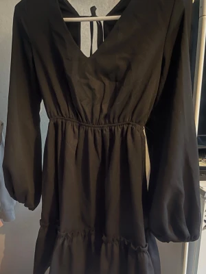 Svart klänning med v-ringning och knyt - Svart klänning med v-ringning och knyt i ryggen. Klänningen har långa ärmar och en volangdetalj vid nederkanten. Den är lätt rynkad i midjan och har en flowy passform som ger en snygg siluett. Perfekt för dig som gillar stilrena och trendiga plagg.