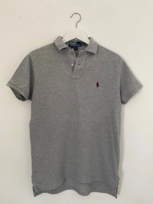 Vintage grå pikétröja från Polo Ralph Lauren - Klassisk grå pikétröja från Polo Ralph Lauren med korta ärmar och krage. Tröjan har två knappar vid halsen och den ikoniska röda logotypen broderad på bröstet. Tillverkad i mjuk bomull för en bekväm och stilren look. Köpt i USA tidigt 2000-tal.