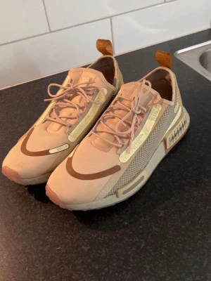 Beige sneakers från Nike med detaljer - Snygga beige sneakers från Nike med unika bruna och ljusgula detaljer. Skorna har meshpaneler på sidorna, textdetaljer och en chunky sula. Perfekta för dig som gillar streetwear och vill sticka ut med din stil.