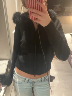 Svart stickad jacka med fuskpäls - Säljer en svart stickad jacka med dragkedja och huva kantad med fuskpäls. Jackan har ribbade muddar och en croppad passform som ger en trendig look. Perfekt för dig som vill ha en mysig men snygg jacka till hösten eller vintern.