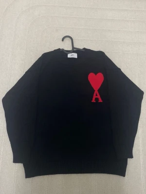 Svart stickad tröja från Ami Paris - Säljer en svart stickad tröja från Ami Paris med deras ikoniska röda hjärta och bokstaven A broderat på bröstet. Tröjan har rund halsringning och ribbade muddar. Perfekt för dig som gillar stilrena plagg med en twist. Storlek M men passar mer S
