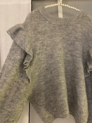 Grå stickad tröja med volangdetaljer - Mysig grå stickad tröja med volanger längs axlarna och ärmarna. Tröjan har rund halsringning och är tillverkad i ett mjukt, ribbstickat material. Perfekt för dig som gillar en trendig och bekväm stil. Passar xs/s/m! 🤗