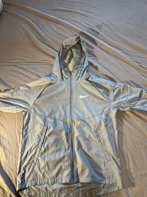 Ljusgrå vindjacka från Nike Running - Snygg ljusgrå vindjacka från Nike Running med huva och hel dragkedja framtill. Jackan har två sidofickor med dragkedja och en minimalistisk Nike-logga på bröstet. Tillverkad i lätt och vindtätt material, perfekt för löpning eller utomhusaktiviteter.