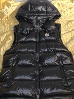 Svart dunväst från Moncler med huva - Snygg svart dunväst från Moncler med glansig finish och avtagbar huva. Västen har två dragkedjefickor framtill och klassisk Moncler-logga på bröstet. Perfekt för lager-på-lager och streetstyle. Insidan har ett unikt serietryck. Helt ny oanvänd äkta.(NFC tag)