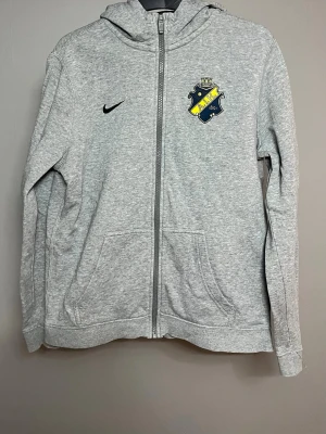 Grå AIK hoodie från Nike - Grå zip-hoodie från Nike med AIK:s klubbmärke på bröstet och svart Nike-logga. Hoodien har huva, dragkedja framtill och två stora fickor. Perfekt för dig som vill visa ditt AIK-stöd med stil. Hör av dig vid funderingar. Priset kan diskuteras vid en snabb affär.🤗🤘🏻