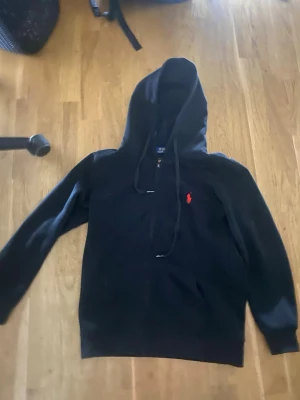 Svart zip hoodie från Polo Ralph Lauren - Klassisk svart hoodie med dragkedja från Polo Ralph Lauren. Har huva med dragsko och den ikoniska röda loggan broderad på bröstet. Tillverkad i mjukt material och har fickor framtill. Perfekt för en avslappnad och stilren look.
