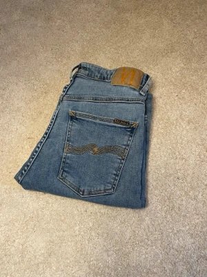 Blå jeans från Nudie Jeans - Säljer ett par klassiska blå jeans från Nudie Jeans med snygg gul söm och vågig brodyr på bakfickan. Jeansen har en traditionell femficksdesign och är gjorda i slitstarkt denim. Perfekta för dig som gillar en tidlös och avslappnad stil.