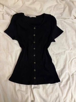 Svart ribbad topp från H&M - Säljer en svart ribbad topp från H&M med korta ärmar och knappar hela vägen framtill. Toppen har en figurnära passform och är tillverkad i mjukt material som känns skönt mot huden. Perfekt att styla med jeans eller kjol.