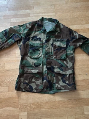 Camouflage overshirt U.S. Marines - Säljer en klassisk militär overshirt från U.S. Marines i camouflage-mönster. Jackan har fyra stora fickor framtill, krage och knäppning. Perfekt för dig som gillar militär saker 