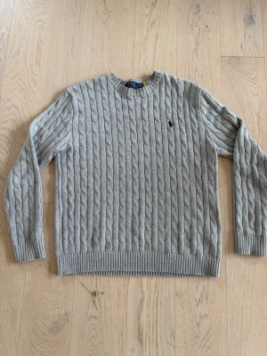Grå kabelstickad tröja Polo Ralph Lauren - Snygg grå kabelstickad tröja från Polo Ralph Lauren med rund halsringning och ribbade muddar. Tröjan har det klassiska broderade logotypen på bröstet och är tillverkad i mjukt material som känns skönt mot huden.