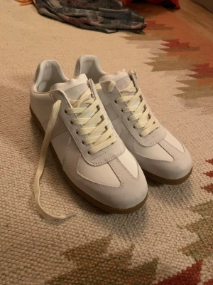 Maison Margiela GATs - Säljer ett par stilrena vita sneakers från Maison Margiela med beige detaljer och klassisk gummisula. Skorna har en låg silhuett, vita skosnören och paneler i både läder och mocka för en clean look. Perfekta för dig som gillar minimalistisk och trendig stil. Innermått: 28,5 