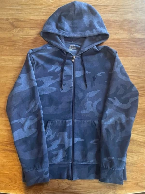 Camouflage hoodie från Polo Ralph Lauren - Polo Ralph Lauren zip hoodie i camo 🔥  Snygg mörkblå kamouflage hoodie från Polo Ralph Lauren med broderad logga på bröstet. Klassisk zip-up modell med luva. Perfekt till både jeans och annat  • Storlek: Medium • Färg: Mörkblå camo • Zip-up • Broderad logo • Skönt och mjukt material  Har ett litet rivmärke men det är diskret och syns knappt när man har på sig tröjan. I övrigt fint skick.  