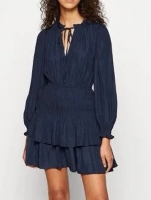 Marinblå volangklänning från maje  - Snygg marinblå kortklänning med långa ärmar och volangdetaljer. Klänningen har smock i midjan, v-ringning med knytband och ett luftigt, flowy tyg.