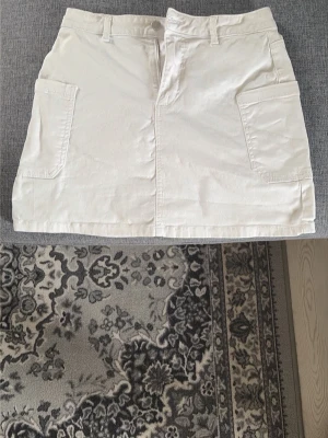 Vit kortkjol från Unique You - Säljer en vit kortkjol från Unique You i denim med stora fickor framtill och dragkedja. Kjolen har bälteshällor och en enkel, clean look som passar till många olika stilar.