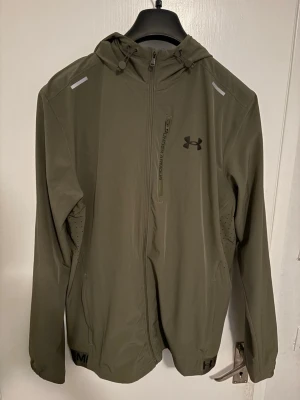 Olivgrön vindjacka från Under Armour - Snygg olivgrön vindjacka från Under Armour med huva, hel dragkedja och bröstficka med logga. Jackan har ventilerande paneler på sidorna och elastisk nederkant med tryckt logotyp. Perfekt för blåsiga dagar och sportiga outfits.