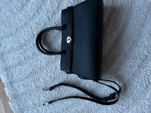 Svart handväska med hjärtspänne - Stilren svart handväska i slätt material med dubbla handtag och avtagbar axelrem. Väskan har ett unikt silverfärgat hjärtformat spänne framtill som detalj. Perfekt för dig som gillar minimalistisk design med en söt twist.