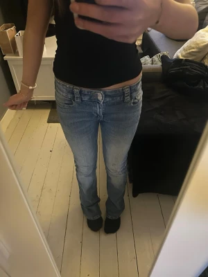 Ljusblå bootcut jeans med ficklock - Säljer ett par ljusblå bootcut jeans med snygg tvätt och ficklock bak med knappdetaljer. Jeansen har låg midja och klassisk femficksmodell. (De ser ut som fläckar på ena benet men de va så när jag köpte dom)