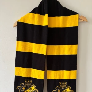 AIK halsduk i svart och gult - Snygg halsduk med breda svarta och gula ränder samt AIK:s klubbmärke broderat i båda ändar. Perfekt för att visa ditt stöd på matchen eller som accessoar till din outfit. Mjuk och skön, troligen i akryl eller liknande material.