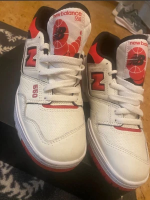 New Balance 550 vita och röda sneakers - Snygga New Balance 550 sneakers i vitt läder med röda och svarta detaljer. Klassisk siluett med perforerade paneler, tydlig branding på plös och häl samt röd yttersula. Perfekta för dig som gillar retrostil och streetwear.