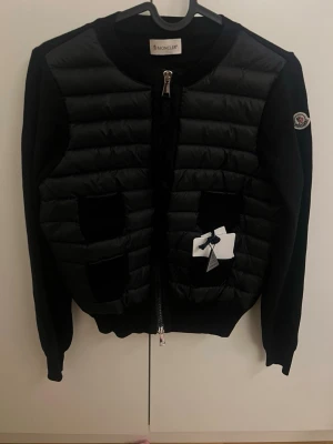 Svart cardiganjacka från Moncler - Snygg svart cardiganjacka från Moncler med quiltad front och mjuka stickade ärmar. Jackan har rund hals, dragkedja framtill och fyra fickor på framsidan. Moncler-logga på ärmen och exklusiv känsla i materialmixen av dun och ull.