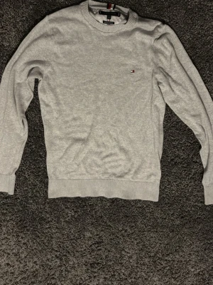Grå sweatshirt från Tommy Hilfiger - Klassisk grå sweatshirt från Tommy Hilfiger med rund halsringning och diskret logga broderad på bröstet. Tröjan har ribbade muddar vid ärmslut och nederkant. Perfekt för en avslappnad och stilren look.