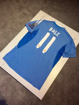 Real Madrid bortatröja Bale #11 - Säljer en blå Real Madrid bortatröja från Adidas med Bale och nummer 11 på ryggen. Tröjan har Champions League-patchar på ärmarna, Fly Emirates-tryck på bröstet och klubbmärke. Tillverkad i funktionsmaterial, perfekt för fotboll eller samling.