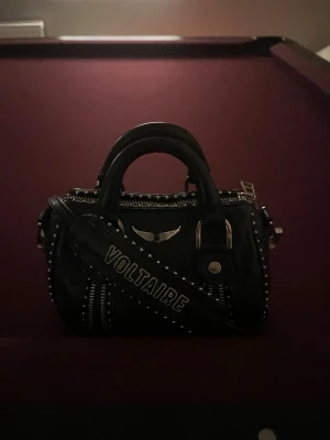 Svart handväska Zadig & Voltaire - Cool svart handväska från Zadig & Voltaire i läder med silverfärgade nitar och detaljer. Väskan har ett brett axelband med texten 'VOLTAIRE', dubbla handtag och dragkedja upptill. Snygg logga med vingar framtill och flera metalldetaljer för en edgy look. I fint skick men har tappat lite färg där på metallen finns på bild men annars i fint skick! Självklart äkta (tillkommer äkta hets bevis och dustbag) Vid snabb affär kan jag tänka mig att gå ner lite i pris! 