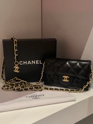  Svart mini väska - Supersnygg svart quiltad handväska insperierad av chanel med klassiskt CC-logo i guld framtill. Väskan har en lång axelrem i kedja med insytt läder och är tillverkad i läder. Perfekt för dig som vill ha en ikonisk och trendig accessoar. Man får inte så mycket plats med saker i väskan pågrund av att den är så liten men kanske mini saker som läppstift samt kort 