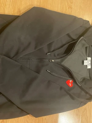 Svart hoodie Comme des Garçons Play - Svart hoodie från Comme des Garçons Play med dragkedja, huva och snörning. På bröstet finns det klassiska röda hjärtat med ögon och på ryggen flera röda hjärtan samt texten 'PLAY COMME des GARÇONS' i vitt. Perfekt för dig som gillar streetwear och snygga detaljer.