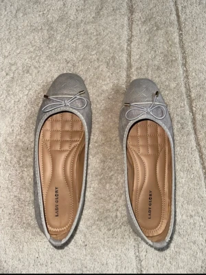 Grå ballerinaskor från Lady Glory - Snygga grå ballerinaskor från Lady Glory med quiltad innersula för extra komfort. Skorna har rund tå, dekorativ rosett framtill och guldfärgade detaljer på snöret. Yttermaterialet är textil med diskret mönster. Perfekta för en stilren look.