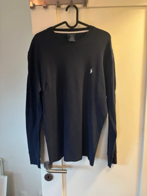 Mörkblå långärmad tröja Polo Ralph Lauren - Snygg mörkblå långärmad tröja från Polo Ralph Lauren med rund halsringning och diskret broderad logga på bröstet. Tillverkad i ett mjukt, strukturerat material som ger en avslappnad men stilren look. Perfekt till jeans eller chinos.