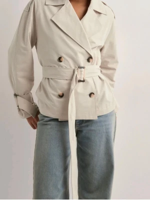 Beige kort trenchcoat med bälte - Trench cost från Nelly storlek xxs, knappast andvänd. Vid frågor och privata bilder är det bara att skriva🫶🏻💓