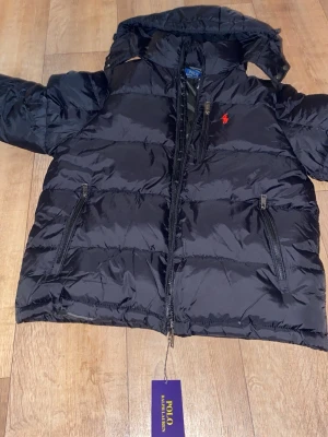 Svart pufferjacka från Polo Ralph Lauren - Svart pufferjacka från Polo Ralph Lauren med hög krage, två dragkedjefickor och klassisk röd logga på bröstet. Jackan har quiltad design och är tillverkad i glansigt material som ger en modern look. Perfekt för kalla dagar.