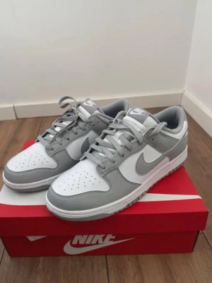 Nike Dunk Low grå och vita sneakers - Nike Dunk Low sneakers i en clean grå och vit färgkombination. Skorna har klassisk låg siluett, perforerad tåbox och ikoniska swoosh-loggan på sidan. Tillverkade i läder med matchande grå snörning och vit mellansula. Perfekta för en stilren streetwear-look.
