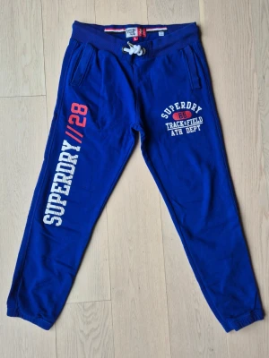 Blå Superdry mjukisbyxor - Snygga blå mjukisbyxor från Superdry med vita och röda tryckdetaljer på benet och logga på höften. Byxorna har dragsko i midjan, två sidofickor och en bakficka. Tillverkade i mjukt bomullsmaterial för maximal komfort.