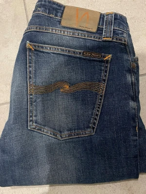 Mörkblå jeans från Nudie Jeans - Snygga mörkblå jeans från Nudie Jeans med klassisk femficksdesign och orangea kontrastsömmar. Jeansen har en smal passform och är tillverkade i denim med lätt slitning för en cool look. Perfekta för dig som gillar stilrena och tidlösa jeans. 