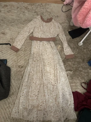 Beige långklänning med blommönster - Elegant beige långklänning med vitt blommönster och matchande bälte i midjan. Materialet känns lätt och flowy, perfekt för vår eller sommar. 