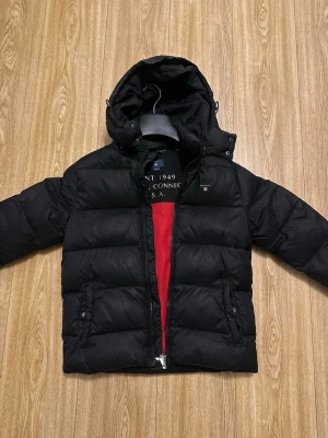 Svart pufferjacka från Gant - Snygg svart pufferjacka från Gant med luva och dragkedja. Jackan har en cool röd insida och praktiska fickor med knappar. Perfekt för kalla dagar och har en klassisk, sportig look. Materialet är vadderat och håller värmen bra.