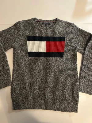 Grå stickad tröja Tommy Hilfiger - Säljer en gråmelerad stickad tröja från Tommy Hilfiger med stort broderat logomärke i vitt, rött och marinblått på bröstet. Tröjan har rund halsringning. 