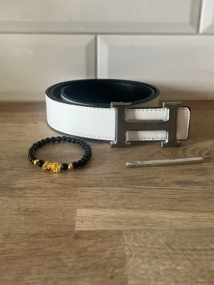 Vitt skärp med silverfärgat H-spänne - Snyggt vitt bälte med ett stort, silverfärgat H-format spänne. Bältet har en stilren design och är gjort i skinn med svart insida. Perfekt accessoar för att lyfta din outfit. Armband ingår!