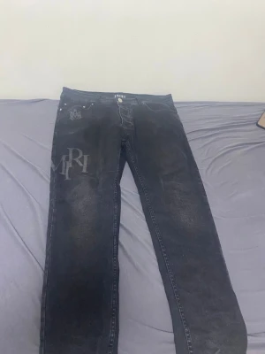 Svarta jeans från Amiri med tryck - Säljer ett par svarta jeans från Amiri med coola tryck på vänster lår och höft. Jeansen har klassisk femficksmodell, bälteshällor och knappgylf. Materialet är denim med en lätt tvättad look och raka ben. Perfekta för dig som gillar streetwear och vill sticka ut.
