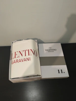 Valentino Garavani vita sneakers - Stilrena vita sneakers från Valentino Garavani med svart band och nitar över vristen. Skorna har klassisk låg profil, rund tå och är tillverkade i slätt läder. Perfekta för dig som vill ha en lyxig och trendig look. Fastpris 