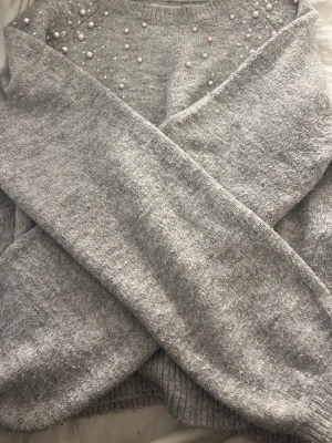 Grå stickad tröja med pärlor - Mysig grå stickad tröja med rund hals och dekorativa pärlor över bröstet och axlarna. Tröjan har långa ärmar och ribbade muddar vid ärmslut och nederkant. Perfekt för dig som gillar en söt och trendig look. Säljs då jag inte använder den och den är knappt använd