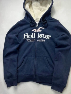 Marinblå Hollister hoodie med foder - Mysig marinblå hoodie från Hollister med dragkedja och vit fluffig teddyfoder på insidan. Stor logga och text på bröstet, två fickor framtill och justerbar huva med snören. Perfekt för kalla dagar och chill vibes.