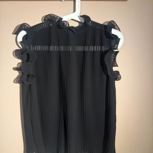 Svart plisserad blus med volang - Snygg svart blus med plisserat tyg och volangdetaljer vid ärmöppningarna och halsen. Blusen är ärmlös och har en lätt transparent känsla upptill, vilket ger en trendig och luftig look. 