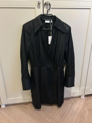 Svart klänning i satin - Stilren svart omlottklänning från Vila med lång ärm och markerad krage. Klänningen har en kort längd och är tillverkad i ett glansigt material som ger en lyxig känsla. Oanvänd med prislapp kvar, normal i storlek. Säljer då fel storlek i present!   500kr i nypris