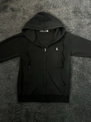 Svart hoodie från Polo Ralph Lauren - Svart hoodie med dragkedja från Polo Ralph Lauren. Klassisk design med huva, dragsnören och två fickor framtill. Liten broderad logga på bröstet. Perfekt för en chill och stilren look.