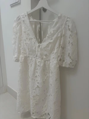 Vit spetsklänning med puffärm - Superfin vit klänning med v-ringning och puffärmar. Klänningen har spetsdetaljer med blommigt mönster och knytning i ryggen. Perfekt för dig som gillar romantisk och feminin stil. Lätt och luftig modell som känns extra somrig.
