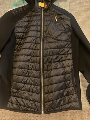 Svart pufferjacka från Parajumpers - Säljer en svart pufferjacka från Parajumpers med huva och dragkedja framtill. Jackan har quiltad front, släta ärmar och en bröstficka med dragkedja. Materialet är glansigt och jackan har en sportig look med detaljer i orange och gult. Fick den som en julklapp men efter bara någon månad utan användning blev den väldigt kort och liten på mig. 